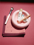 ILIA makeup products including a brush, complexion stick, lip pencil, sharpener, and lip + cheek stick on a pink plate and scarf.