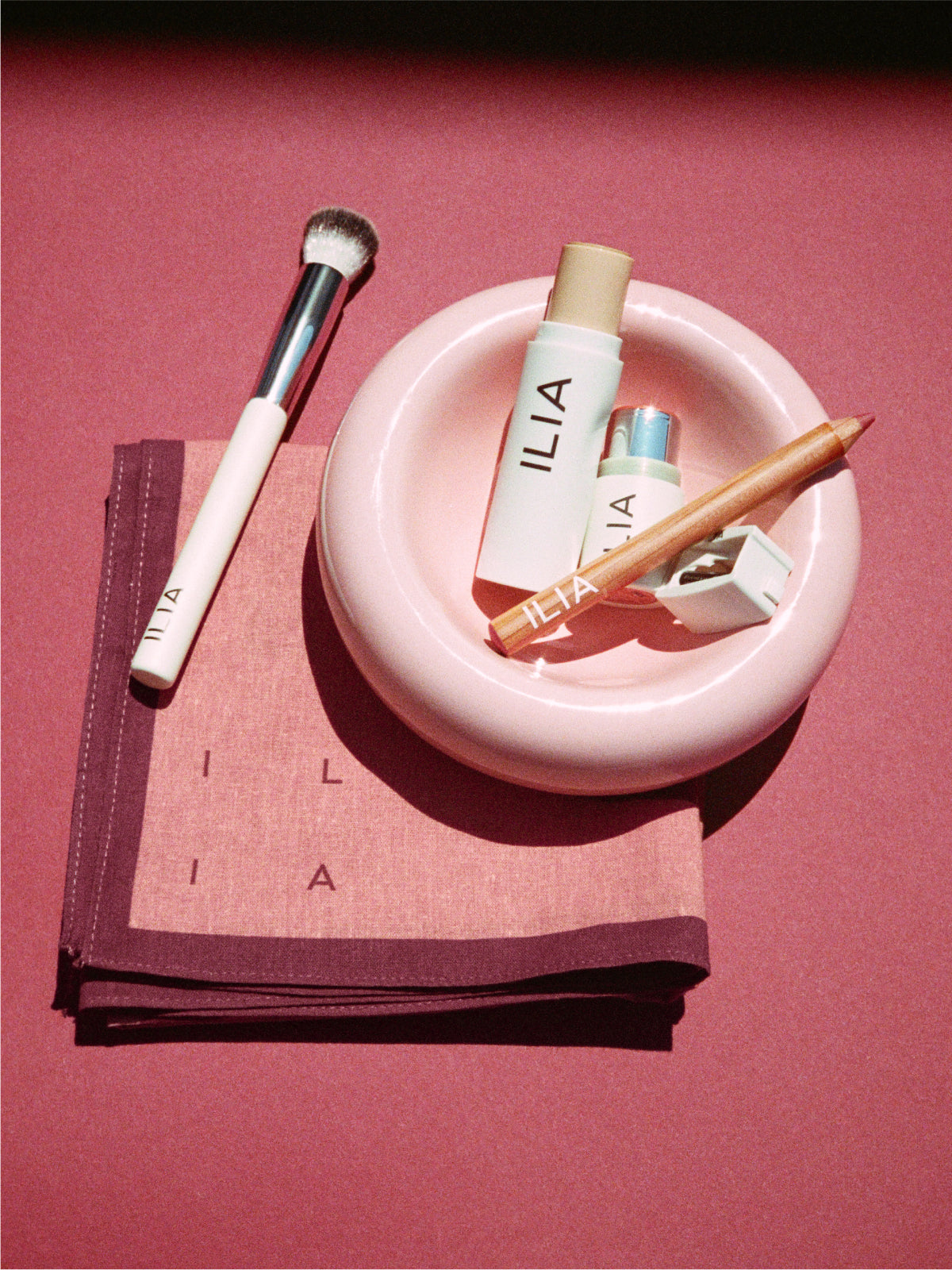 ILIA makeup products including a brush, complexion stick, lip pencil, sharpener, and lip + cheek stick on a pink plate and scarf.